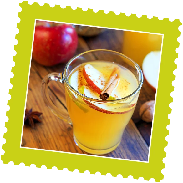 A glass of warm apple cider with cinnamon and apple slices, surrounded by fresh apples and spices on a wooden table.