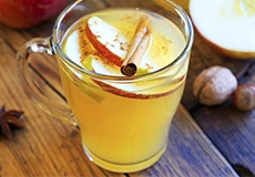 A glass mug filled with hot apple cider, garnished with apple slices and a cinnamon stick, sits on a wooden surface.