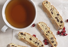 A cup of tea sits next to four pieces of biscotti and a few pomegranate seeds on a white surface.