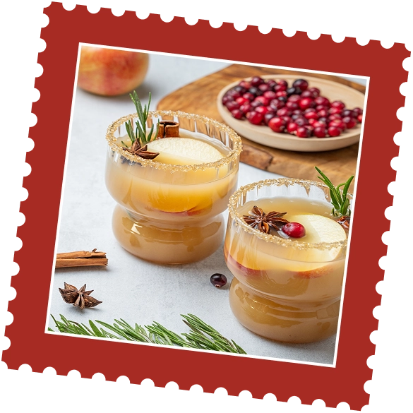 Two glasses of spiced apple cider garnished with apple slices, cranberries, rosemary, and star anise, with cranberries and spices visible in the background.
