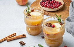 Two glasses of apple cider garnished with apple slices, cranberries, rosemary, and star anise, with cinnamon sticks and a bowl of cranberries nearby.