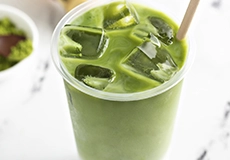 A clear plastic cup filled with iced matcha green tea latte and a straw, placed on a white surface.