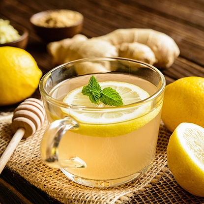 A glass cup of lemon ginger tea garnished with a lemon slice and mint, surrounded by fresh lemons, ginger root, a honey dipper, and a bowl of honey on a rustic surface.