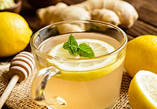 A glass mug of lemon ginger tea garnished with a mint sprig, surrounded by fresh lemons, ginger root, and a honey dipper on a rustic surface.